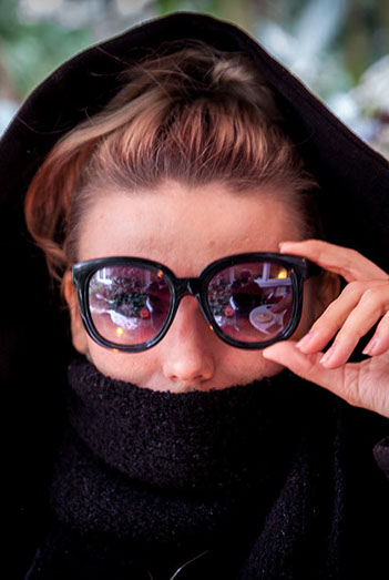 Arina legt Ihre Hand auf Ihre Sonnenbrille und schaut zur vierten Wand.   