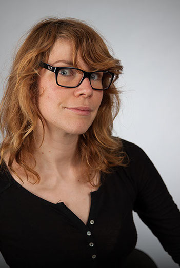 Adrienn mit RotBlonden Haaren, Brille mit Schwarzen Gestell und Schwarzen Oberteil steht im Fotostudio vor einer Grauenfotowand.    